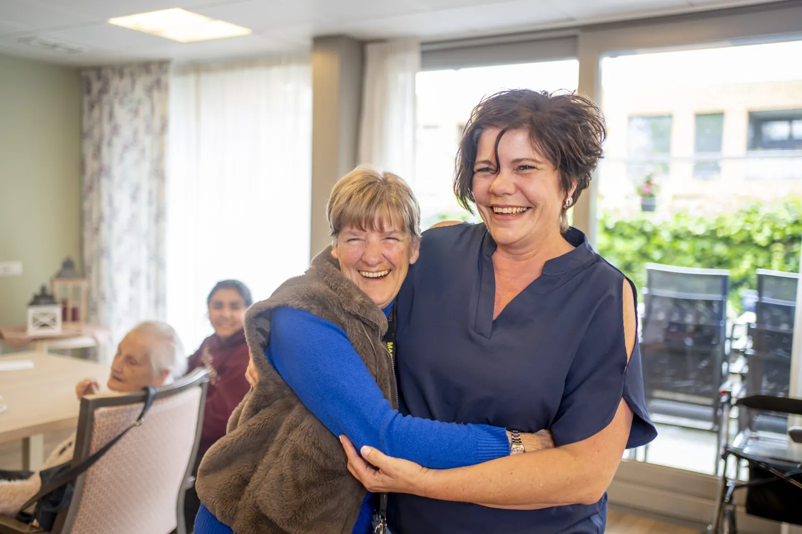 Twee vrouwen omhelzen elkaar lachend in een huiskamer van Liemerije