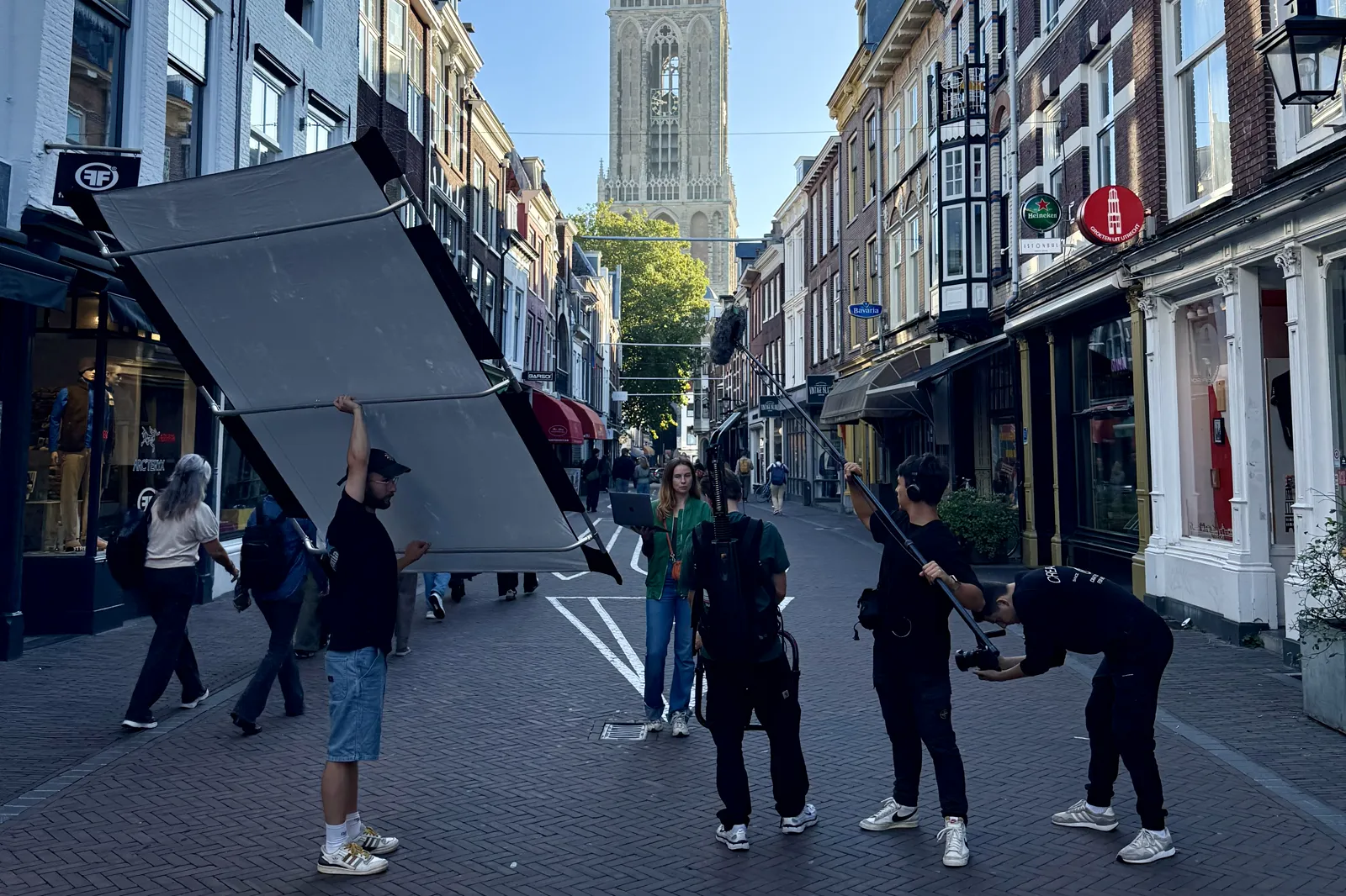 De crew draait in een winkelstraat in Utrecht met de Domtoren op de achtergrond: een man tilt een groot scherm, anderen houden microfoon en camera