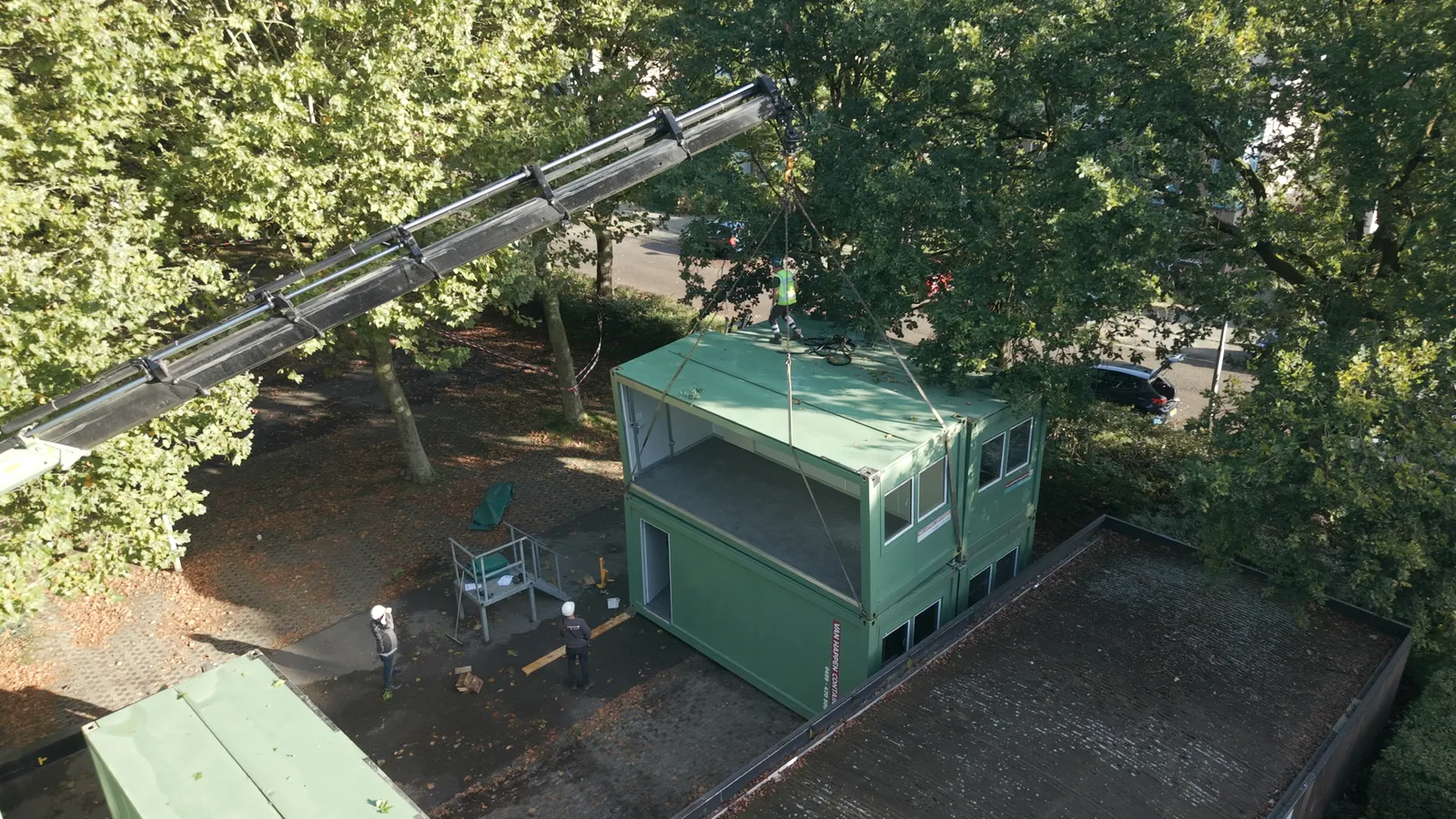 Dronebeeld van een kraan die een groene bouwunit op een andere plaatst, tussen hoge bomen, met bouwvakkers ernaast